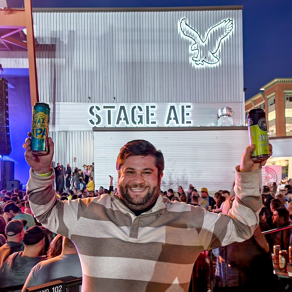 Alex at Stage AE in front of the Stage AE sign holding two beers