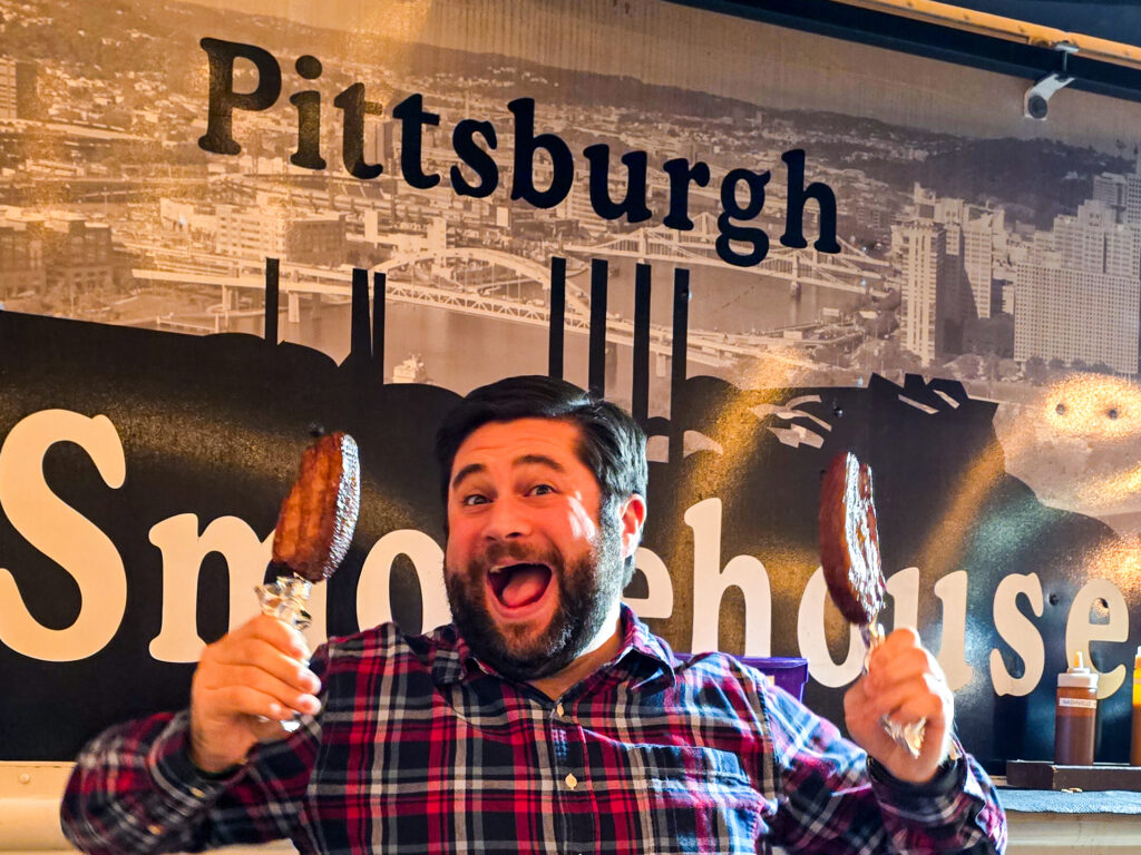 alex in front of the pittsburgh smokehouse food truck holding two bacon on a stick