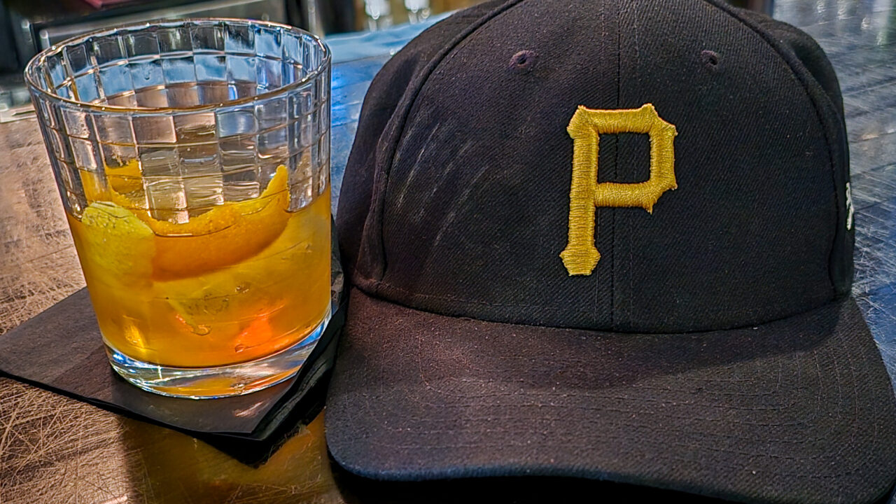 Fred's Old Fashioned Cocktail from The Warren on the left next to a black Pittsburgh Pirates baseball hat with the yellow Pirates P logo.