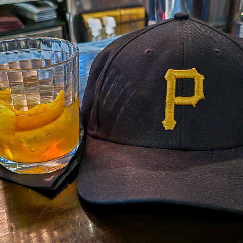 Fred's Old Fashioned Cocktail from The Warren on the left next to a black Pittsburgh Pirates baseball hat with the yellow Pirates P logo.
