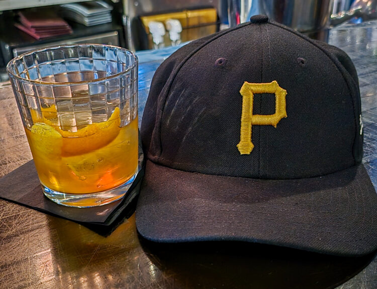 Fred's Old Fashioned Cocktail from The Warren on the left next to a black Pittsburgh Pirates baseball hat with the yellow Pirates P logo.