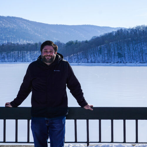 Alex standing in front of a railing at Raystown Dam in Huntingdon, PA in winter with the lake frozen.