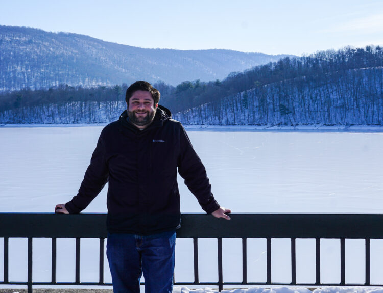 Alex standing in front of a railing at Raystown Dam in Huntingdon, PA in winter with the lake frozen.