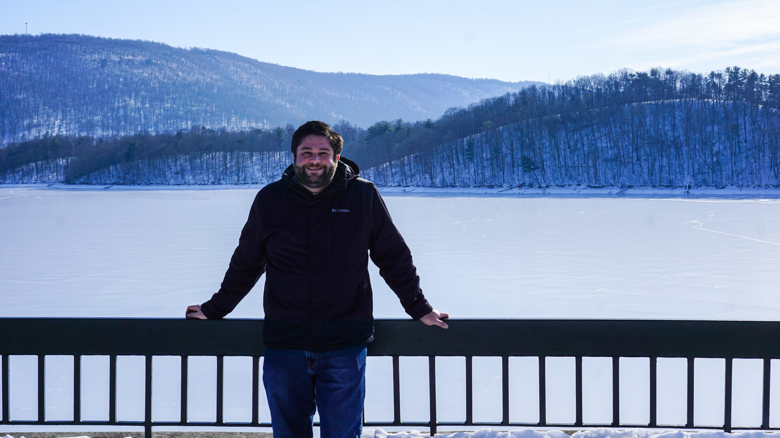 Alex standing in front of a railing at Raystown Dam in Huntingdon, PA in winter with the lake frozen.