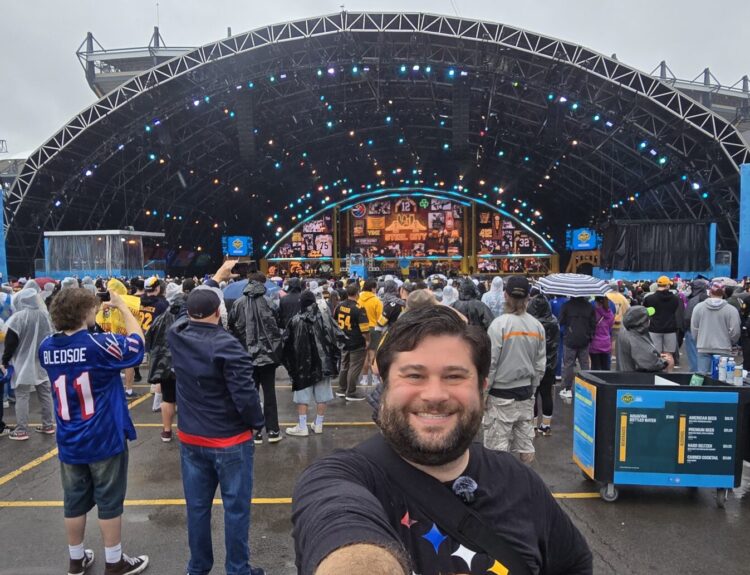 Alex taking a selfie on day three of the 2026 NFL Draft in Pittsburgh. Alex is standing in front of the NFL Draft Stage and it is raining.