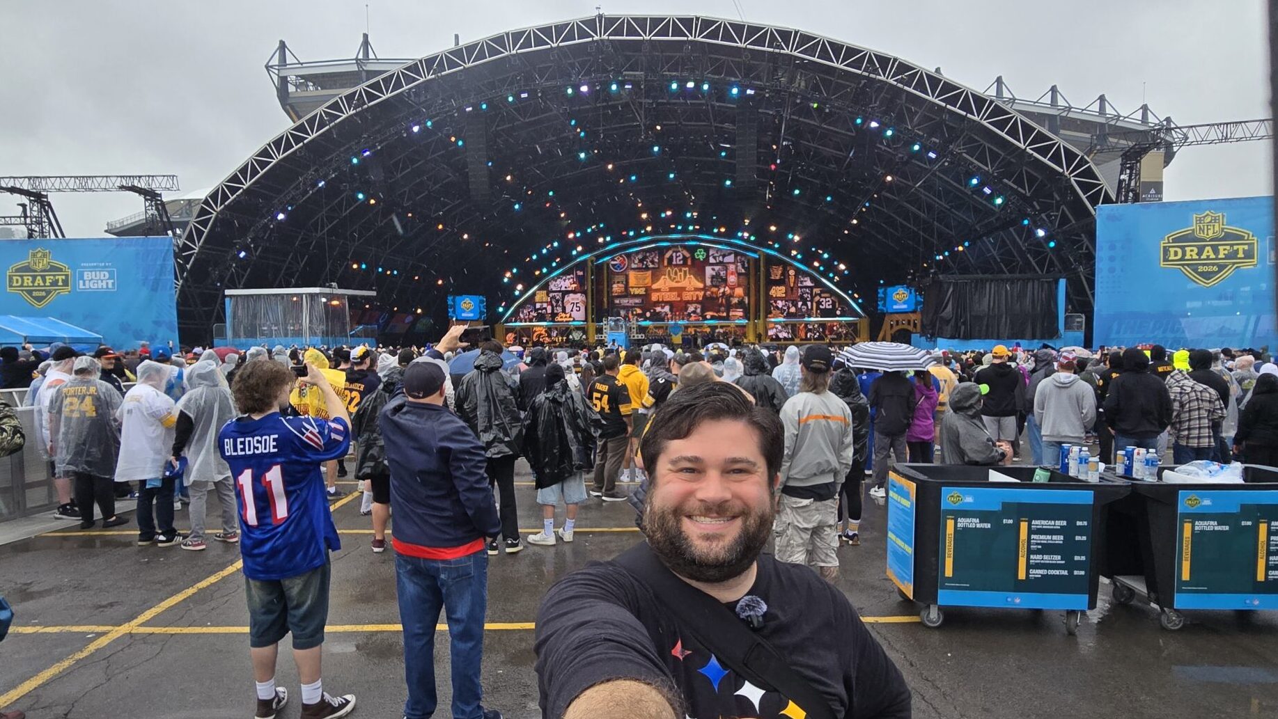 Alex taking a selfie on day three of the 2026 NFL Draft in Pittsburgh. Alex is standing in front of the NFL Draft Stage and it is raining.