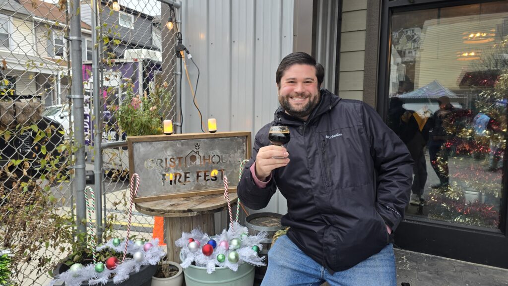 Alex posing in front of a Grist House Fire Fest Metal sign, holding a glass of stout beer.