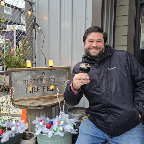 Alex posing in front of a Grist House Fire Fest Metal sign, holding a glass of stout beer.
