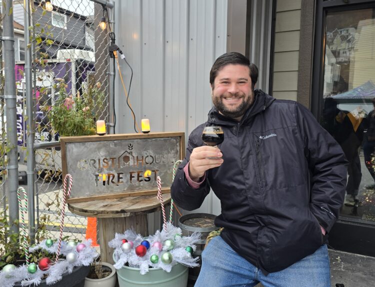 Alex posing in front of a Grist House Fire Fest Metal sign, holding a glass of stout beer.