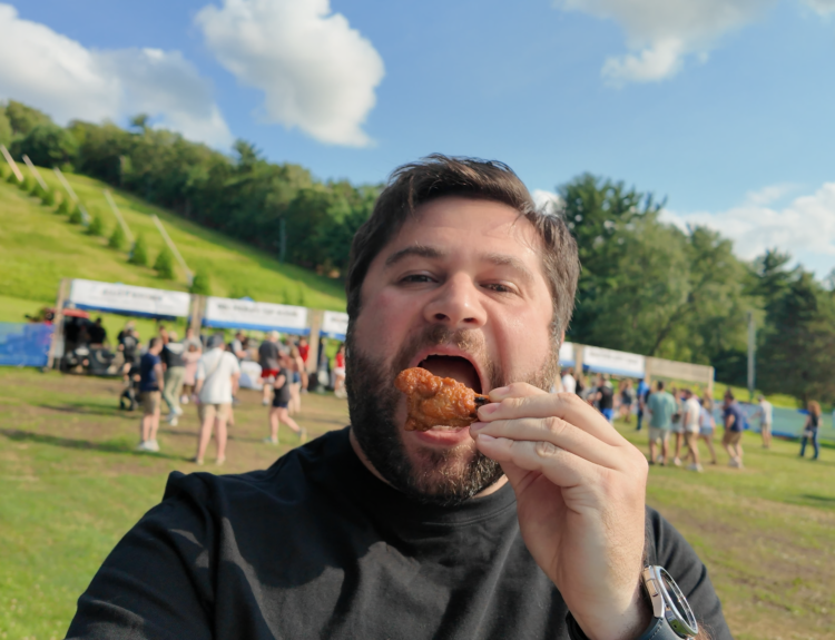 Alex at Tussey Mountain WingFest eating a chicken wing drumstick with the vendor booths in the background